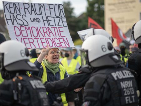 Auf einer Demonstration hält eine Person ein Schild: "Demokratie verteidigen" während sie von Polizisten umzingelt ist. Symbolbild für mögliche Polizeigewalt