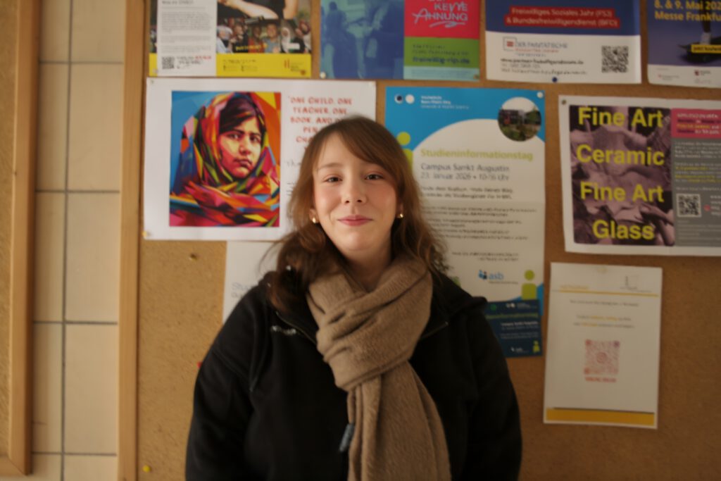 Eine junge Frau mit braunem Schal steht vor einer bunten Pinnwand im Flur von Koblenz-Kolleg.