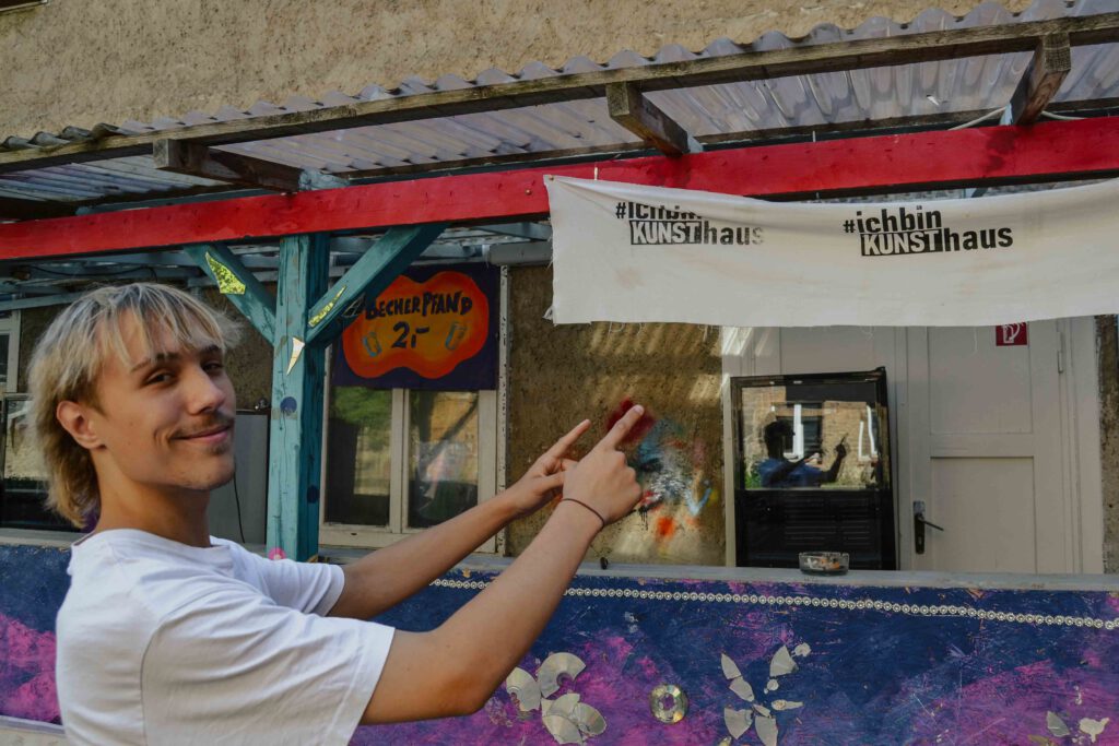 Luca, ein Junge mit blonden, mittellangen Haaren, zeigt auf ein weißes Banner, auf dem „#ich bin KUNSThaus“ steht. Es hängt an einem bunten, überdachten Tresen. 
