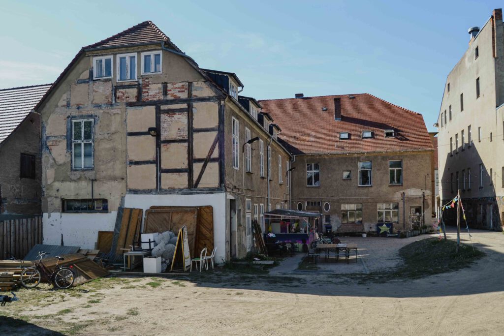 : Das Kunsthaus ist wie ein „L“ angeordnet. Es hat eine grau verputzte Fassade mit Fachwerk am Giebel. An der Seite liegt ein Stapel Holzplatten. In der Mitte stehen Bierbänke an einem bunten Ausschank. Rechts spannt sich eine Girlande über den Hof. 