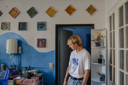 Luca, ein Junge mit blonden, mittellangen Haaren, trägt Jeans und ein weißes T-Shirt. Er steht in der Tür eines Kunstraums mit blau-weißen Wänden. In einem Regal stehen Becher mit Pinseln. An der Wand hängen bunt gestaltete Keramikfliesen.