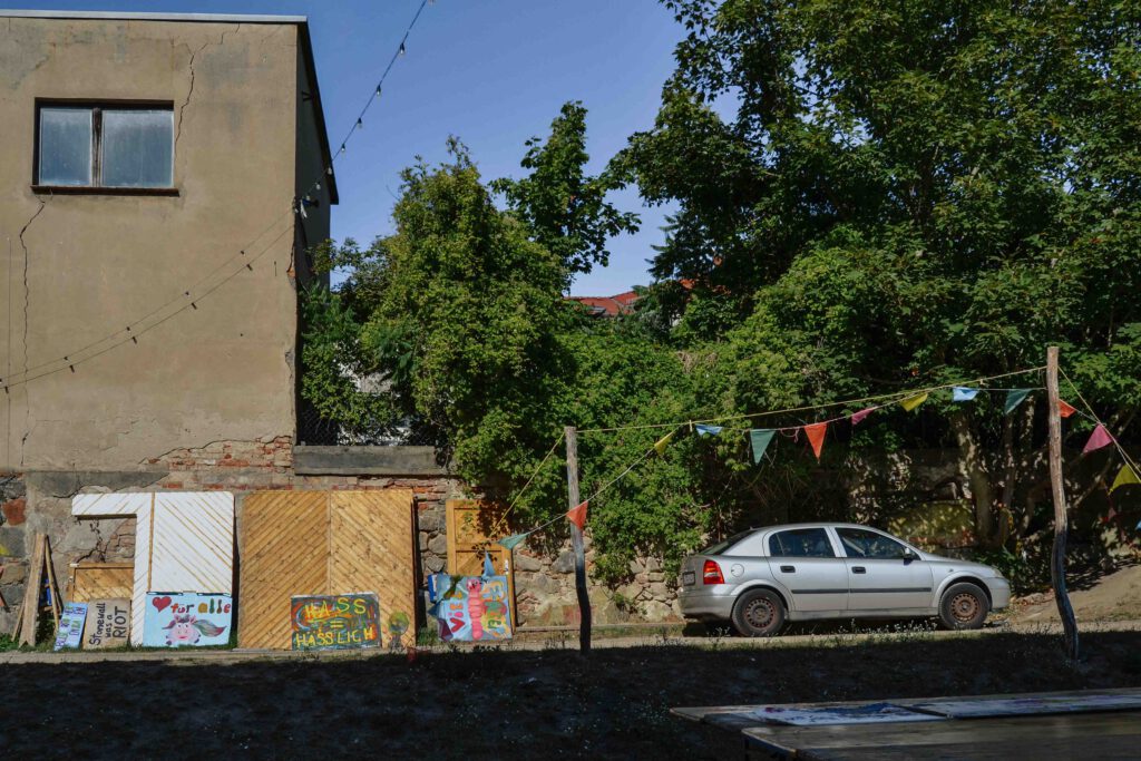 Im Innenhof des Kunsthauses lehnen bunte Plakate mit Aufschriften wie „Hass = hässlich“ oder „<3 für alle“ an einer Wand mit bröckelndem Putz. Girlanden spannen sich über den Hof. Rechts steht ein silbernes Auto unter Bäumen. 