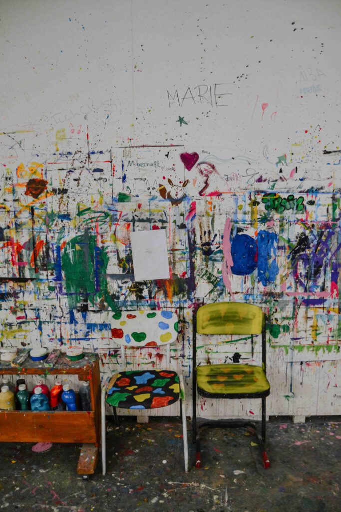 Eine Wand ist unregelmäßig mit bunter Farbe bemalt. Vereinzelt sind Namen und Symbole zu erkennen. Davor stehen zwei bunt bemalte Stühle und ein Holzregal mit Farbtuben. 